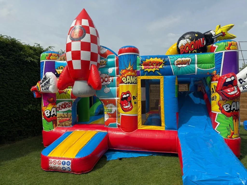 Het kleurrijke Multiplay Comic springkasteel met raket en glijbaan op een zonnig grasveld